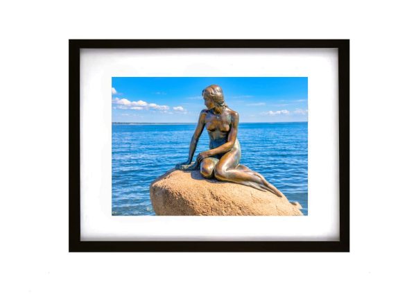 A bronze statue of a mermaid sitting on a rock in the water at Langelinie pier, Copenhagen, under a bright blue sky.